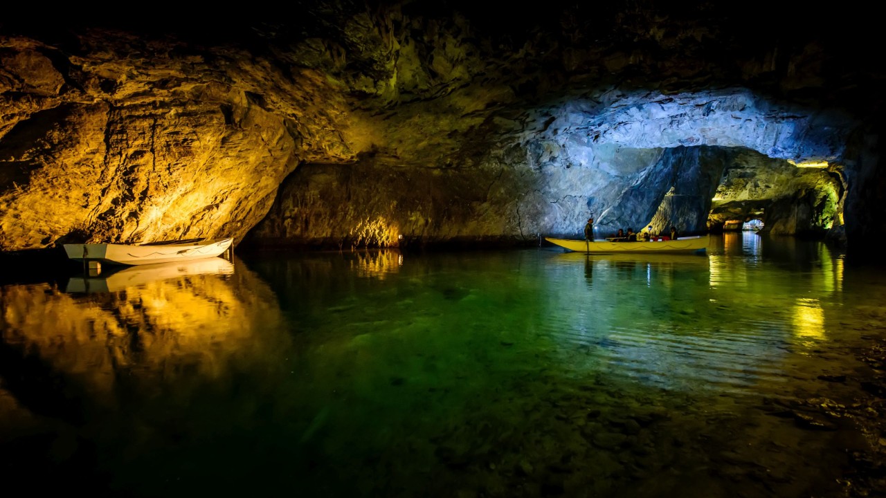 Das Gewölbe einer Grotte spiegelt sich im Halbdunkel im kristallklaren Wasser eines unterirdischen Sees. Im Hintergrund gleitet ein Boot durch einen engen Gang, der eine Verlängerung der Grotte bildet.